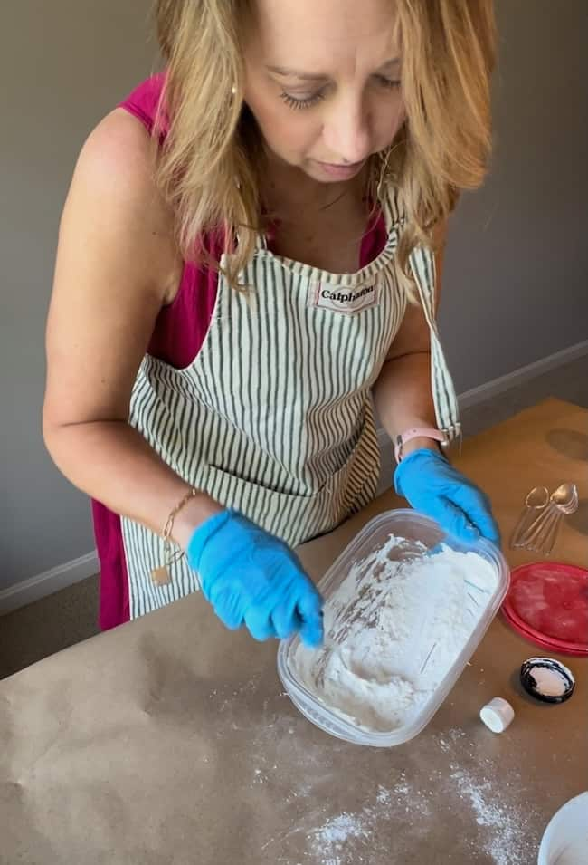 A woman with light brown hair is wearing a striped apron and blue gloves. She is mixing a white substance in a plastic container on a table covered with brown paper, next to measuring spoons and a red container lid. An Anthropologie-inspired jute twine vase adds charm to the scene.