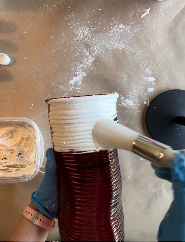 A pair of hands wearing blue gloves are applying white plaster to the upper part of a dark red vase using a brush. A plastic container with white plaster sits nearby on a table covered in brown paper with white powder scattered around.