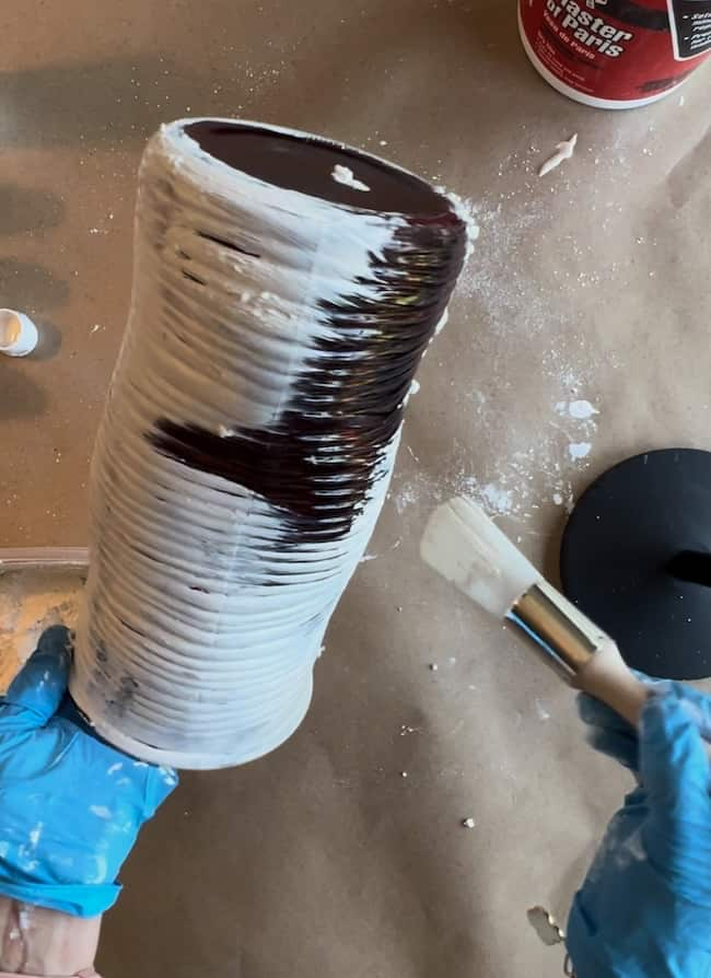 A person wearing blue gloves is applying white plaster to a tall, ridged vase using a paintbrush. The workspace has evidence of plaster splatters, and a container of plaster is visible in the background.