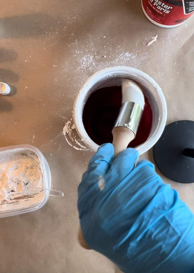 A person wearing a blue glove dips a white paintbrush into a container of dark red liquid. Nearby is an open tub with white material inside and a black lid. A surface is lightly dusted with white powder.