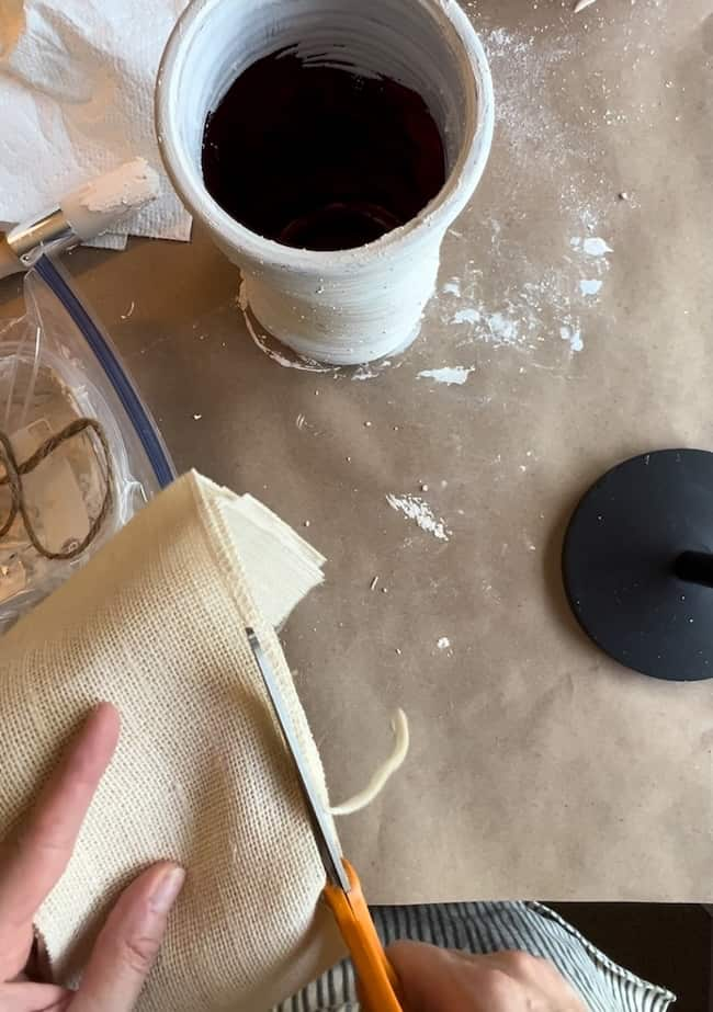 A person is cutting a piece of beige fabric with orange-handled scissors. Nearby, there is a white ceramic container with a dark liquid inside, as well as a spool of jute twine and various crafting materials on a brown surface.