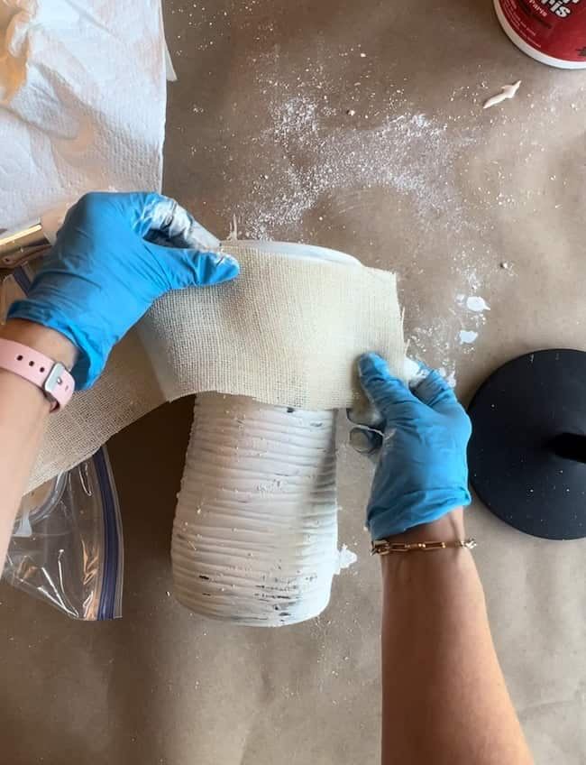 Two hands wearing blue gloves are wrapping a white cylindrical object, possibly a vase, with a piece of plaster cloth. The scene is on a brown work surface with some scattered white plaster powder, a black circular base, and a container in the background.