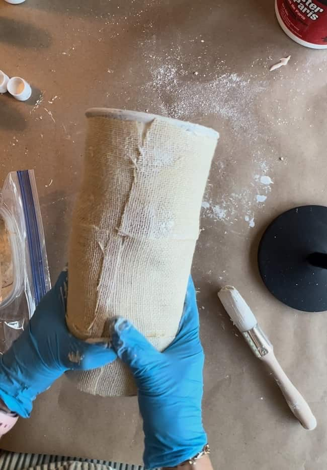 A person wearing blue gloves is holding a cylindrical object wrapped in plaster cloth. A paintbrush and container are nearby on a workspace covered with brown paper and scattered white powder.