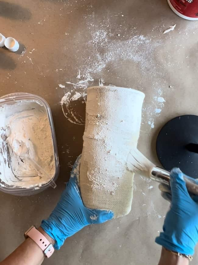 A person wearing blue gloves and a pink wristwatch applies plaster to a cylindrical object, possibly a vase, using a spatula. A container with plaster mixture is visible on the left side, and a round black object is positioned on the right.