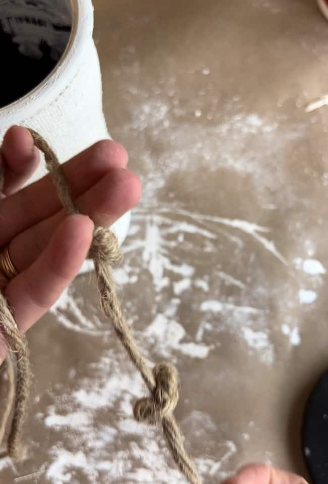 A person's hand is holding a piece of twine with two knots in it. The background includes a white container and a brown surface with white powdery residue scattered on it.