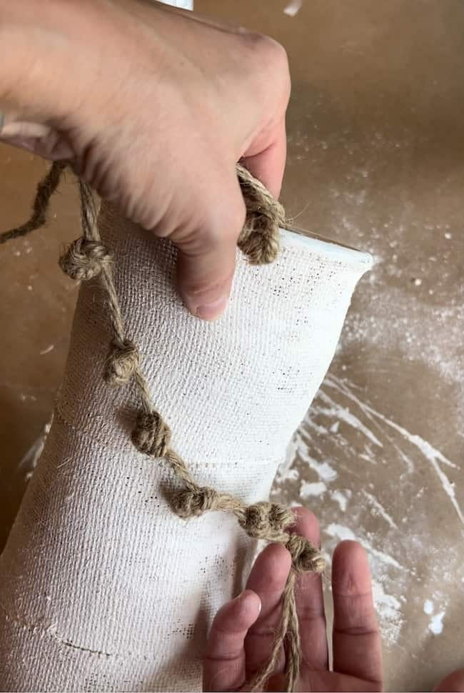 Two hands working on a craft project, holding a piece of burlap or coarse fabric with a knotted rope. The background shows a brown surface with scattered white powder, suggesting an artistic workspace.