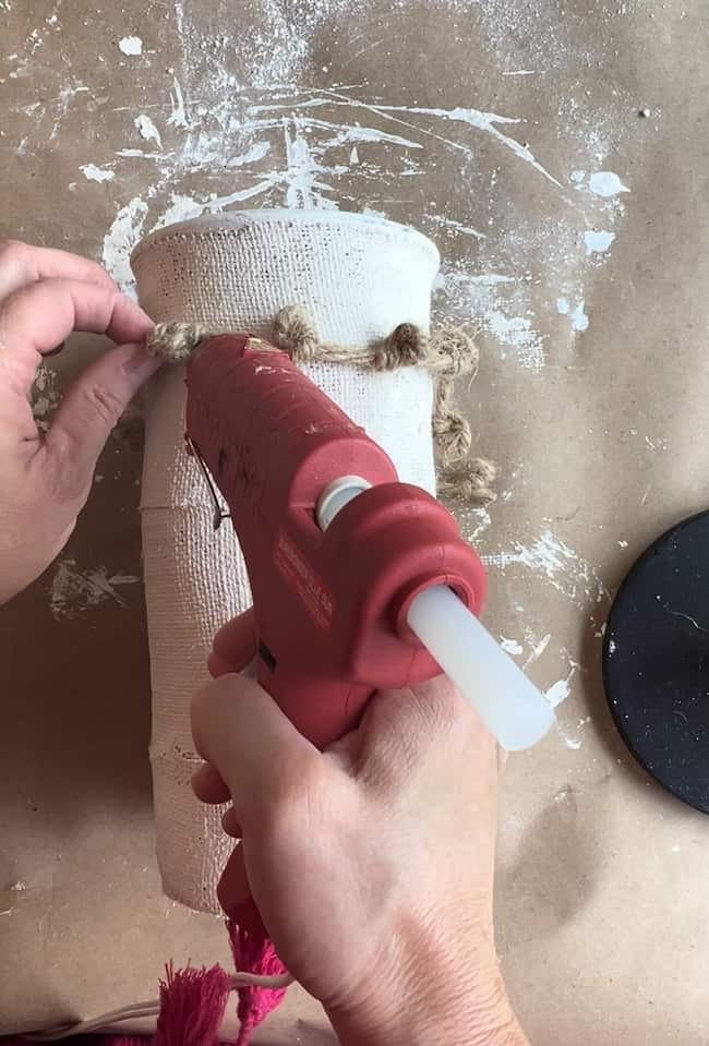 A person uses a red hot glue gun to attach a piece of rope to a cylindrical white object, which appears to be covered in plaster or a similar material. The workspace is covered with brown paper, and white paint or plaster is splattered around.