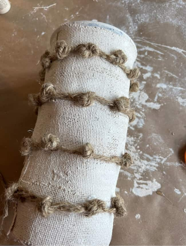 A cylindrical object wrapped in beige fabric with rough, textured jute twine tied around it in evenly spaced horizontal rows. The Anthropologie inspired vase sits against a messy workspace background, cluttered with scattered paint or plaster smudges.