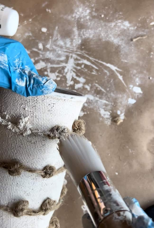 Person wearing a blue glove uses a white paintbrush to apply paint to a textured, white surface with rope accents. The background shows scattered white paint and a brown surface, indicating an ongoing art or DIY project.