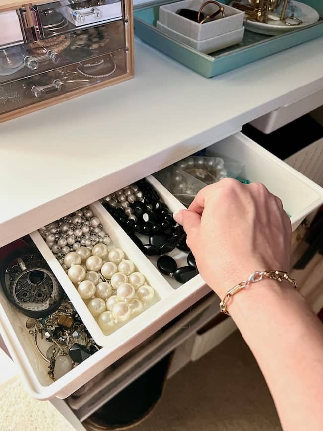 A person's hand is seen opening a meticulously arranged drawer that showcases back-to-school organization ideas for home, with different compartments holding various jewelry items, including pearl beads, black beads, and other trinkets. A jewelry box with more items is on the table above the drawer.