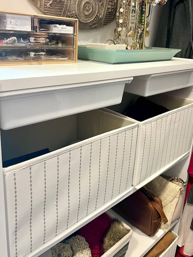 A white shelving unit with pull-out fabric drawers offers perfect back-to-school organization ideas at home. The top shelf holds a jewelry display with assorted necklaces and a box of bracelets. The middle shelf features two striped fabric bins, while the bottom shelf contains folded knitwear and leather handbags.