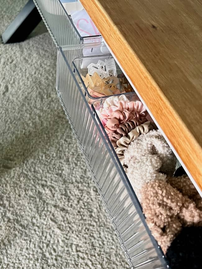 A close-up of an organized drawer with clear dividers containing hair accessories, including scrunchies, hair ties, and clips. Perfect for back to school organization ideas for home, this drawer is under a wooden tabletop, and the surrounding area features a light-colored textured carpet.