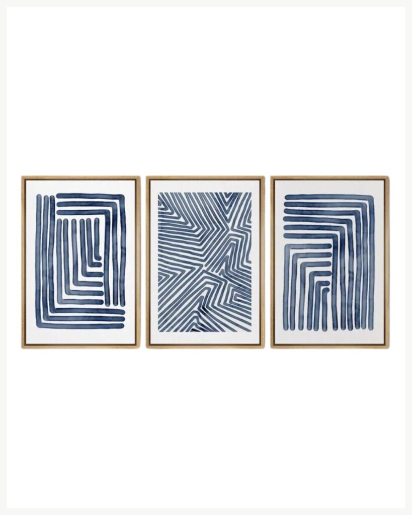 A set of three framed abstract geometric art prints that warm up your space. Each print features varying designs of navy blue parallel lines creating different shapes and patterns. The light-colored wooden frames enhance the cozy living room decor, displayed against a plain white background.