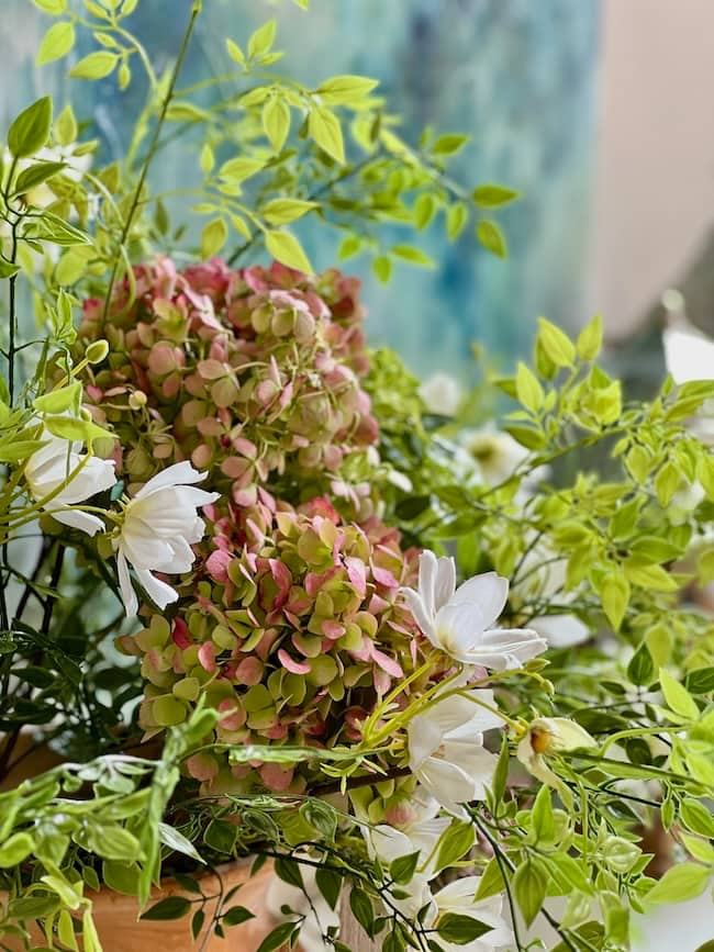 A lush bouquet featuring green and pink hydrangeas, with white flowers interspersed, is surrounded by abundant green foliage. The background is a soft, blurred blend of blue and green hues, making it perfect for cozy living room decor.