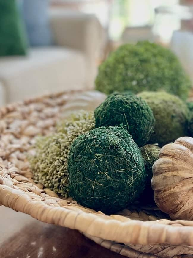 A woven basket filled with decorative green moss balls and an artificial pumpkin sits on a wooden surface. The background shows blurred furniture, suggesting a cozy indoor setting—a perfect touch of cozy living room decor to warm up your space.