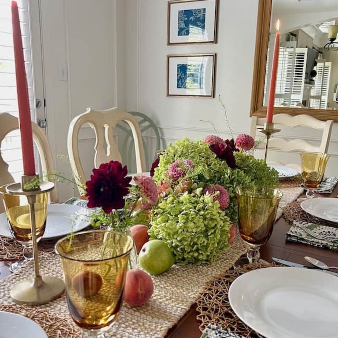 A dining table set for a meal with a centerpiece of lush green hydrangeas, deep red flowers, apples, and peaches showcases end of summer decorating ideas. The table features gold candlesticks with red candles, amber glasses, and white plates on woven placemats. Two framed pictures hang on the wall.