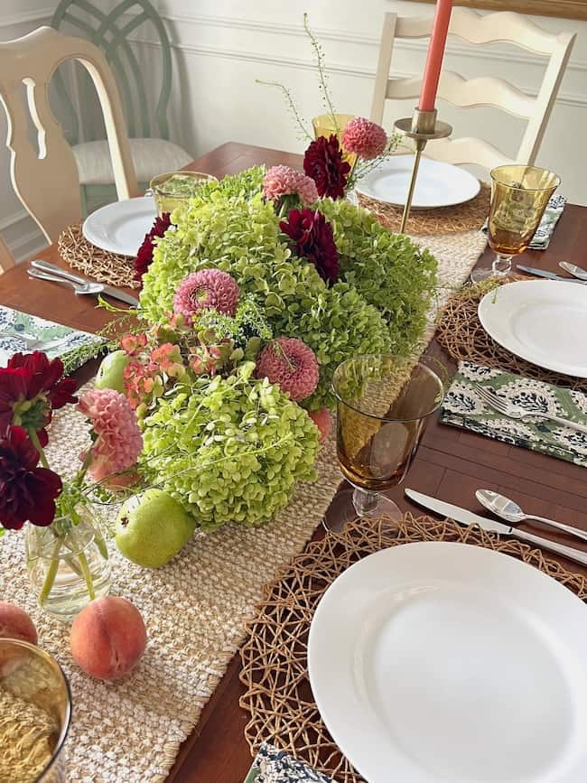 A wooden dining table is set with white plates, amber glasses, and woven placemats. The centerpiece features hydrangea-inspired colors with green hydrangeas, pink and red flowers, green apples, and a peach. A single red candle in a gold holder completes this perfect end-of-summer decorating idea.