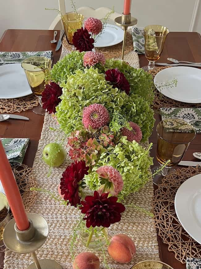 A dining table is decorated with a centerpiece of green hydrangeas and pink and red flowers. Gold candle holders with orange candles, woven placemats, and green drink glasses brighten up your space. Fruits, including apples and peaches, are placed sporadically.