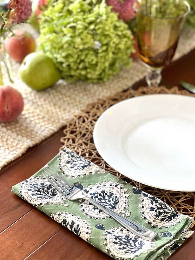 A close-up of a set dinner table with a white plate on a green, patterned napkin featuring a fork. The table decor includes a woven placemat and a brown-rimmed glass. In the background, End-of-Summer Decorating is highlighted with hydrangea-inspired colors - lush green and pink hydrangeas, a green apple, and red peaches.