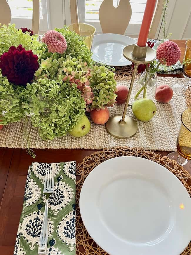 A decorated dining table featuring a green and beige checkered table runner, hydrangea-inspired colors in fresh flowers and fruits, white plates on wicker placemats, green patterned napkins with silver cutlery, amber glasses, and a gold candlestick with a pink candle—perfect to brighten up your space.