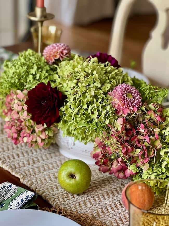 An elegant floral centerpiece with green and pink hydrangeas, dark maroon and pink dahlias, on a woven table runner brightens up your space. A green apple is nearby, along with a lit candle in a brass holder and a glass with a red apple in the background.