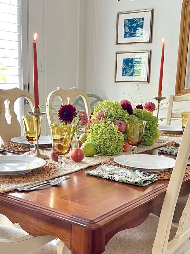 A dining table set for four with a centerpiece of lush green and burgundy flowers, inspired by the hydrangea colors, surrounded by fruits. The table setting includes woven placemats, white plates, amber goblets, and patterned napkins. Two red candles in elegant holders are lit at each end of the table.