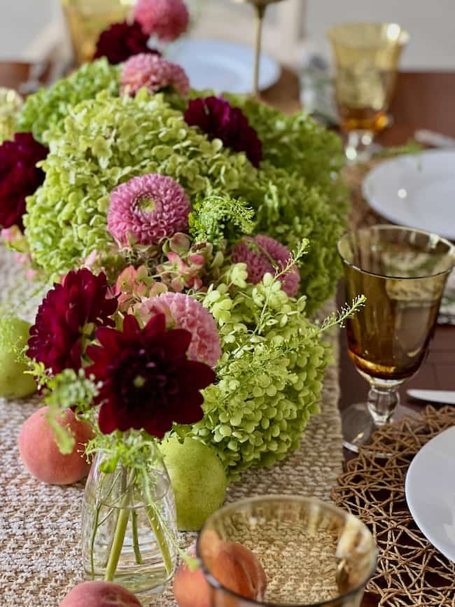 A dining table, perfect for end-of-summer decorating, is adorned with a floral centerpiece featuring green hydrangeas, deep burgundy dahlias, and pink flowers. The table is set with amber-colored glassware, white plates, and woven placemats. Peaches and green pears are scattered decoratively to brighten up your space.