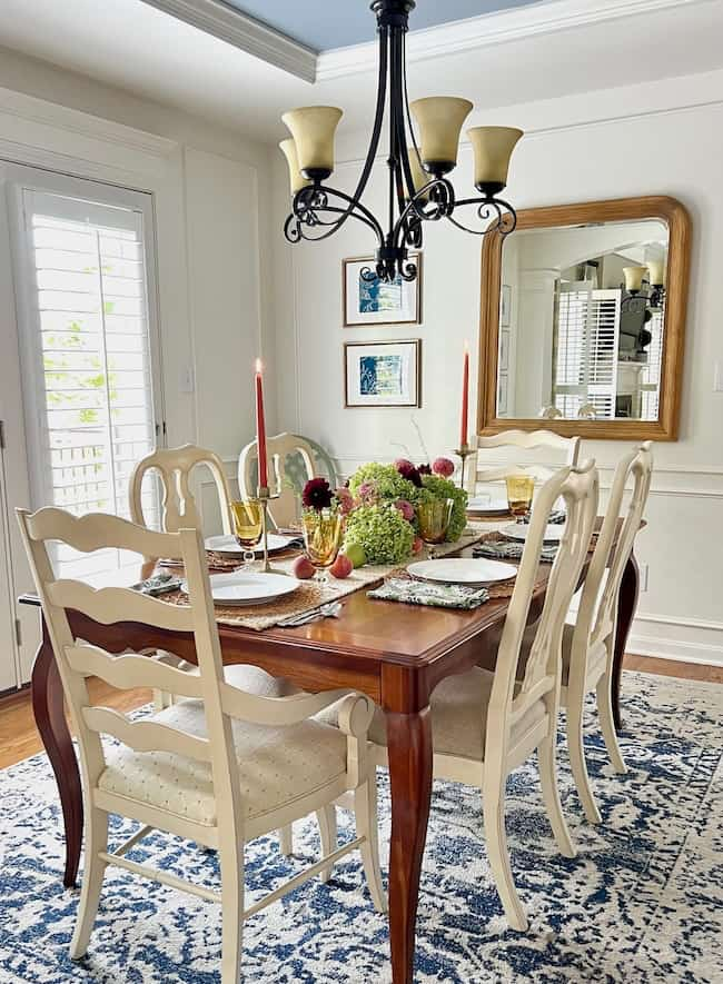 A dining room with a wooden table set for a meal with white chairs around it. The table, adorned with floral centerpieces in hydrangea-inspired colors, candles, and dinnerware, captures the essence of end-of-summer decorating. A large mirror on the wall and a five-light chandelier brighten up your space effortlessly.