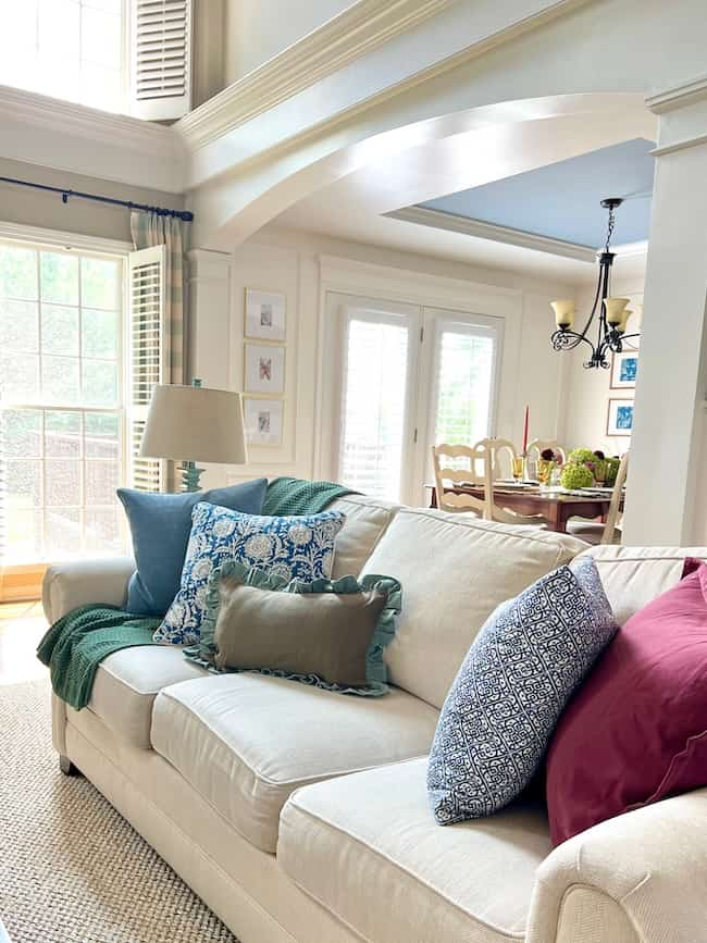 A cozy living room features a beige sofa adorned with an assortment of colorful pillows, inspired by hydrangea pastel hues in blue, green, and red. Overhead, natural light floods in through large windows, brightening up the space. A dining area with a table and chairs is visible in the background.