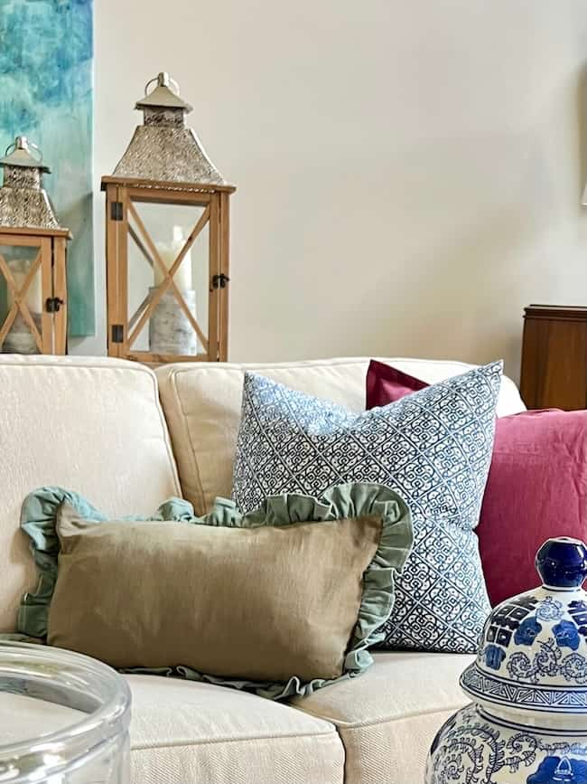 A beige couch adorned with decorative pillows, including a ruffled sage green one, a large patterned blue pillow, and a magenta pillow. Behind the couch are two wooden lanterns with metal tops. An ornate blue and white jar sits on a nearby table. Perfect for end-of-summer decorating!