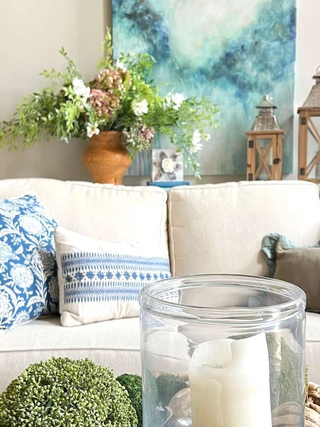 A cozy living room with a beige sofa adorned with blue and white patterned pillows, inspired by end-of-summer decorating. Behind the sofa is a large abstract painting in hydrangea-inspired colors. A glass vase with a candle and green decorative moss sits on the table, brightening up your space with flowers and lanterns in the background.