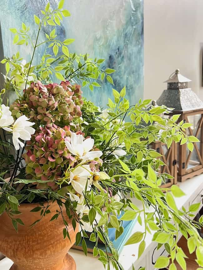 A floral arrangement in a terra cotta pot features green leaves, white flowers, and pink-tinged hydrangeas, perfect for end-of-summer decorating. Behind it, there is a wooden lantern with a metal top and a textured blue and green painting on a white wall.