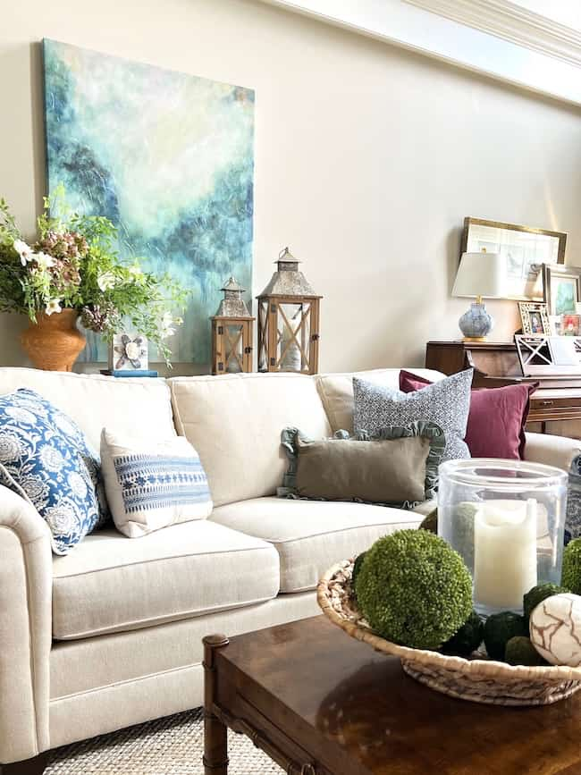 A cozy living room with a beige sofa adorned with various patterned and colored pillows. A table in front of the sofa holds a decorative basket with greenery and candles. The wall behind features a large abstract painting, lanterns, and additional decor that showcases end of summer decorating ideas.