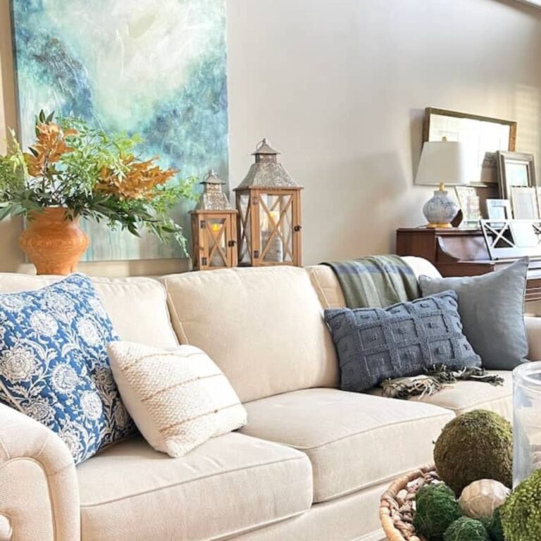 A cozy living room features a beige sofa adorned with various patterned cushions. Embracing fall decorating trends, there are decorative lanterns and a painting with green and blue hues on the wall. A table lamp and a collection of books are placed on a wooden console behind the couch.