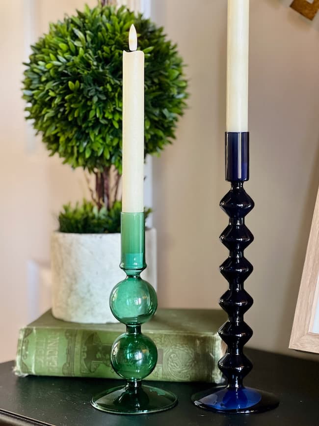 Two candlesticks with lit candles are displayed on a surface. The left candlestick is green and has a spherical design, while the right candlestick is black with a geometric pattern. A potted plant and two green hardcover books are in the background, showcasing affordable Target home decor for Fall Decorating Trends 2024.