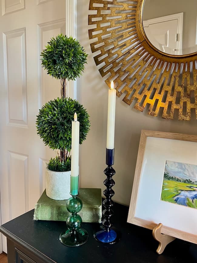 A stylish interior arrangement featuring a small topiary plant, two lit candlesticks in green and black holders on a dark table, and a round, geometric gold mirror hanging above. A framed painting adds charm while reflecting affordable home decor trends. A door is partially visible in the background.