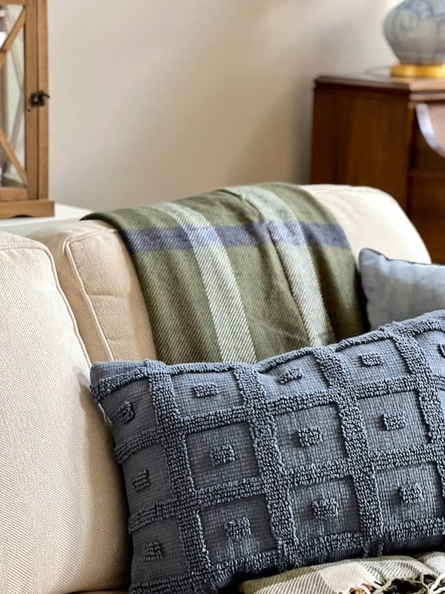 A cozy beige couch with a textured blue-gray pillow and a green plaid throw blanket draped over the back. The scene, inspired by fall decorating trends, is set in a warmly lit room with wooden furniture and Target finds as decorative elements in the background.