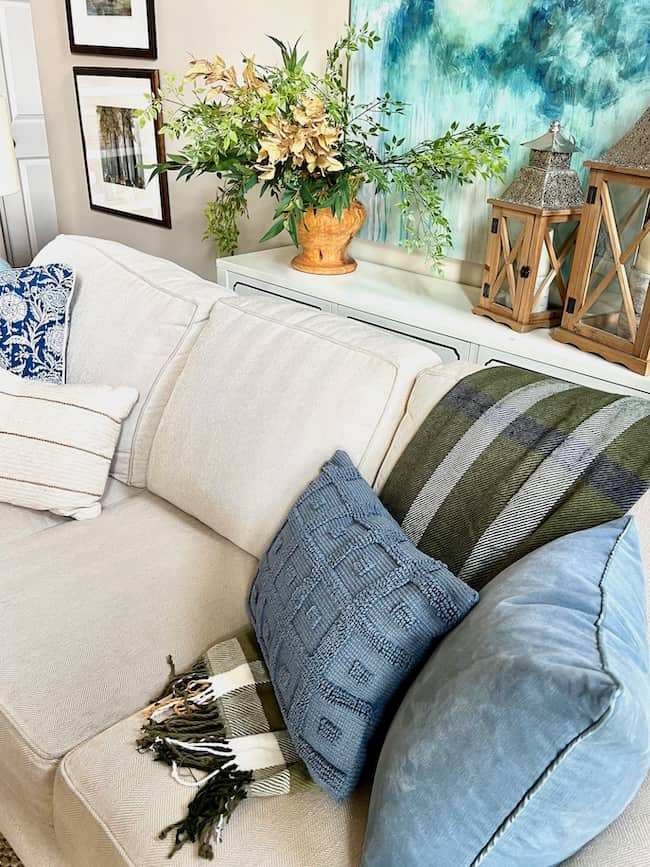 A cozy living room setup featuring a beige couch adorned with various decorative pillows in green, blue, and beige tones. A green and white plaid throw blanket is draped over the armrest, perfect for fall decorating trends. Above, there is a console table with plants, lanterns, and an abstract painting—all stylish Target finds.