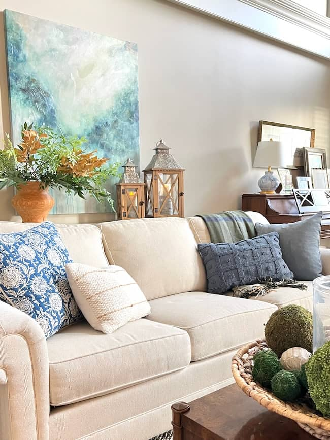 A cozy living room with a beige sofa adorned with blue, gray, and white pillows. Behind the sofa, there are lanterns, a vase of greenery, and a vibrant abstract painting on the wall. Embracing Fall Decorating Trends 2024, a bowl with decorative green spheres from Affordable Target Home Decor sits on the wooden coffee table.