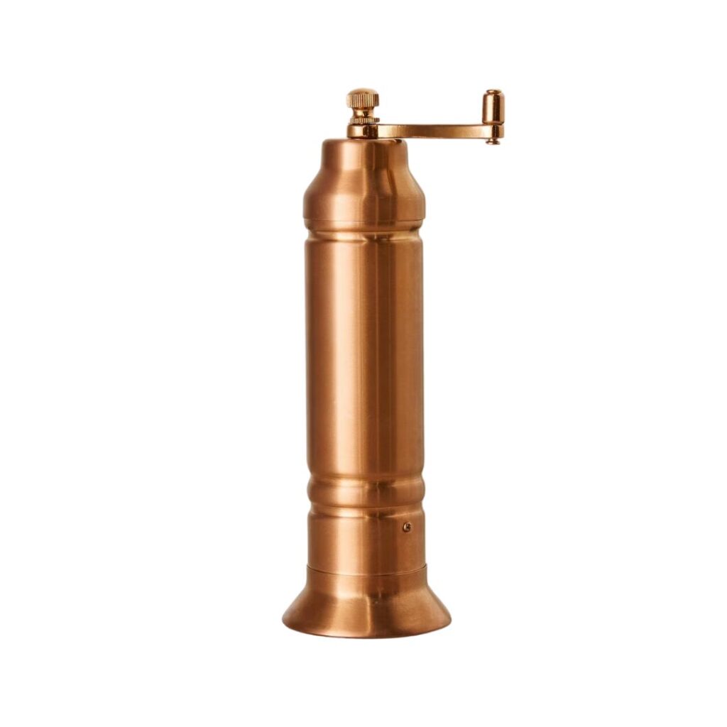 A cylindrical copper pepper mill with a shiny finish features a long handle on top for grinding. The base is slightly wider and flares out. The overall design is simple and elegant, suitable for a kitchen or dining setting. The background is white.