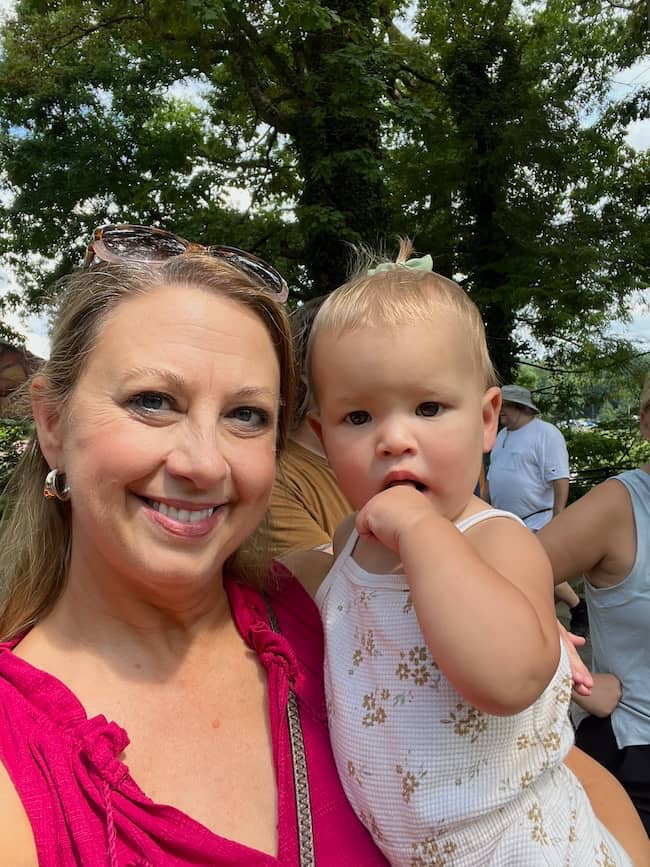 A woman in a pink sleeveless top holds a young child wearing a light-colored floral outfit. The child has a hand near their mouth. They are outdoors with trees and other people in the background. The woman is smiling, and both are looking at the camera.