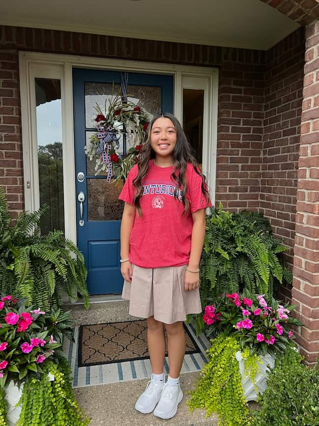 A young girl with long dark hair is standing in front of a house with a brick exterior. She is wearing a red "CENTAURUS" T-shirt, a beige skirt, and white sneakers. There are potted plants with pink flowers and green foliage flanking the entryway.