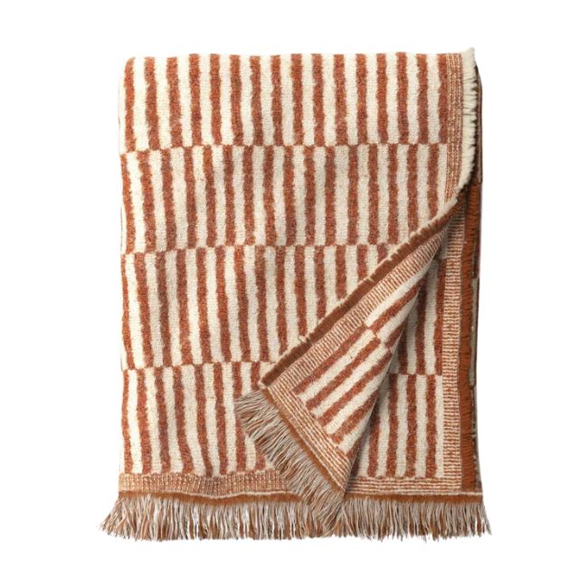 A folded, woven blanket with a pattern of alternating vertical and horizontal rust-colored stripes on a cream background. The blanket features fringed edges and looks soft and cozy.