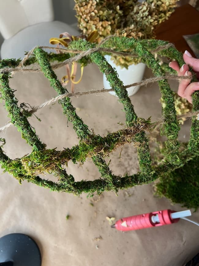 A hand is holding a partially covered spherical frame made of twine, resembling a Dollar Tree pumpkin wreath DIY project. Patches of moss are attached. A hot glue gun and glue sticks are on the table, along with some dried plant material in the background, indicating a craft project in progress.