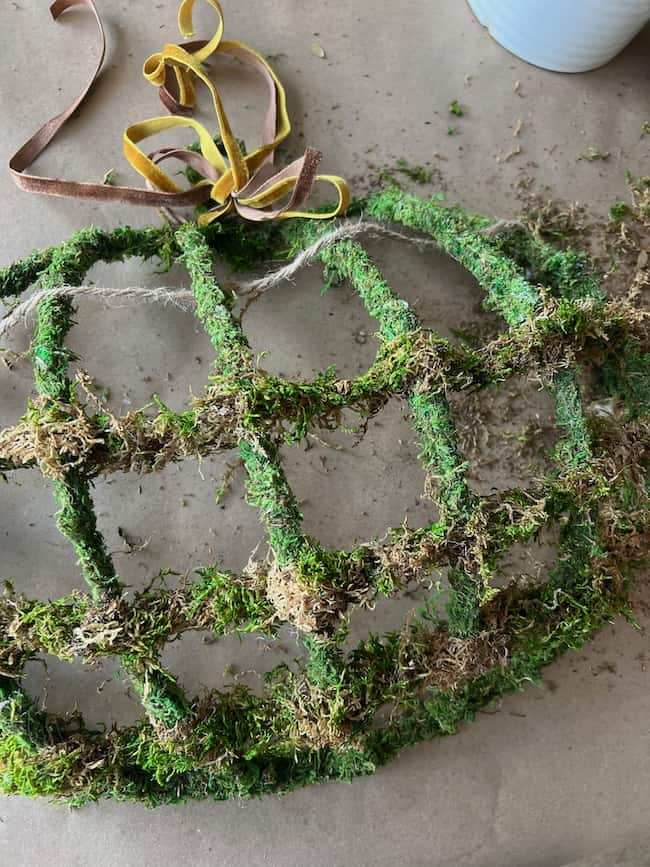 A round, wire frame wrapped with green moss, partially covered in natural fibers, resembles a charming Dollar Tree pumpkin wreath DIY. Two brown ribbons are tied at the top. Moss and plant debris are scattered on the surface around the frame, indicating a crafting or gardening project in progress.