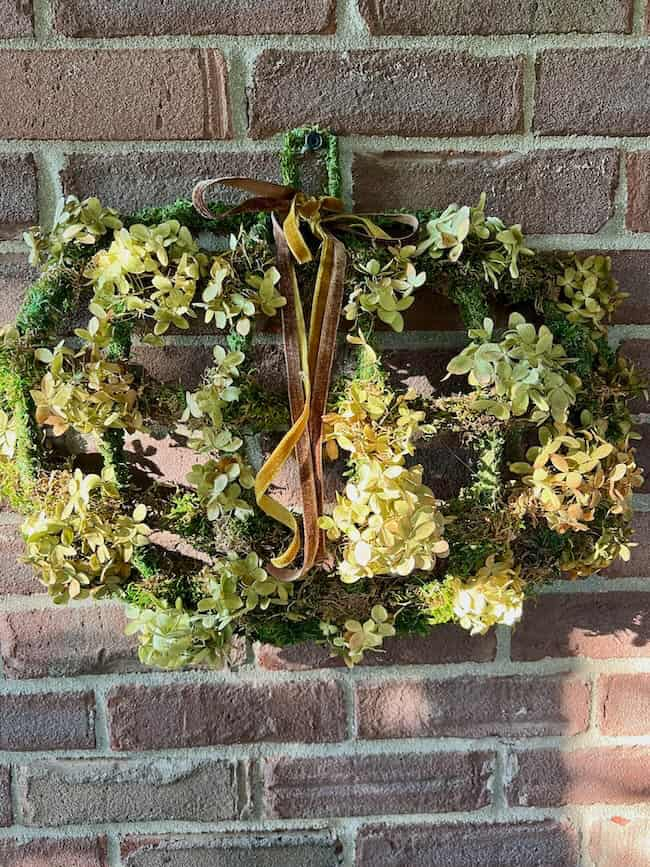 A brick wall features a fall-themed Dollar Tree pumpkin wreath DIY project shaped like a pumpkin. The wreath is adorned with clusters of dried yellow flowers and green moss, accented by a brown ribbon. Sunlight casts shadows across the wall and wreath, highlighting its handcrafted beauty.