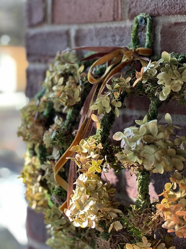 Close-up of a decorative Dollar Tree pumpkin wreath hanging on a brick wall. The wreath is adorned with dried flowers and green foliage, and features a series of ribbon bows in earthy tones. The background is softly blurred, suggesting an outdoor or sunlit setting.