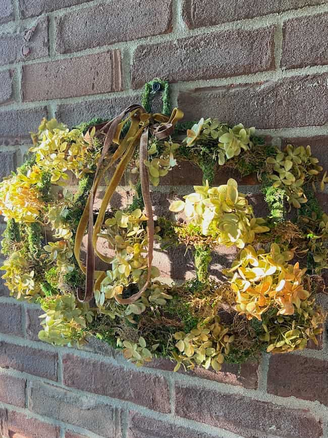 A Dollar Tree pumpkin wreath made of dried and green foliage adorned with brown ribbons hangs on a brick wall. Featuring a mix of yellow and green leaves, it creates a natural and rustic appearance. Sunlight illuminates the wreath, casting shadows on the wall behind it.