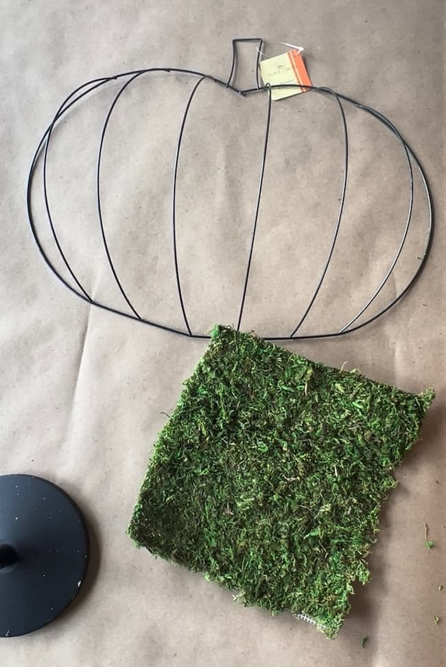 A metal wire frame shaped like a pumpkin and a green square piece of artificial turf or moss are lying on a brown surface. The frame has a small tag attached to its top, perfect for your Dollar Tree pumpkin wreath DIY project.