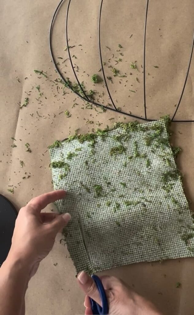 A pair of hands cuts a piece of green, textured fabric with blue-handled scissors. The fabric has small green clippings scattered on it. On the brown surface below are a black circular wire frame—perfect for a Dollar Tree pumpkin wreath DIY—and additional green clippings.